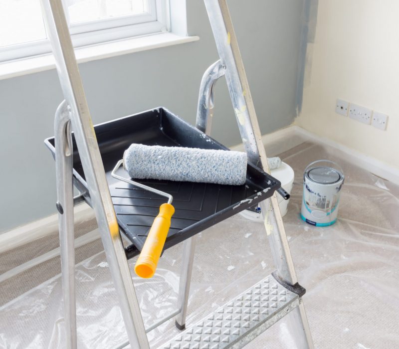 Painting and decorating with a roller and tray on a set of metal step ladders with cans of paint and a dust sheet in the background