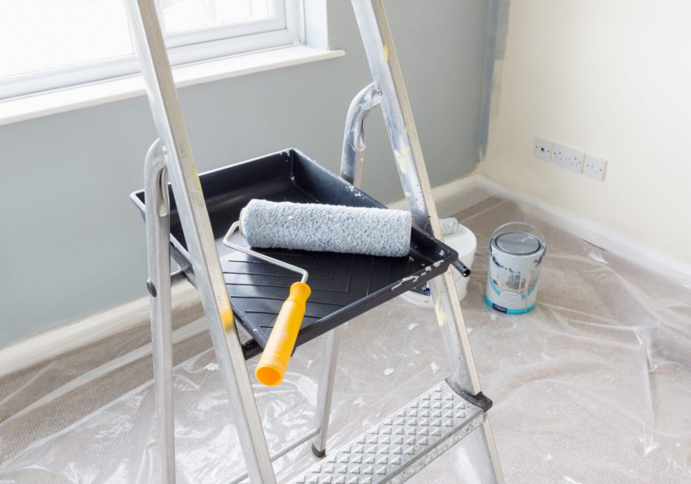 Painting and decorating with a roller and tray on a set of metal step ladders with cans of paint and a dust sheet in the background